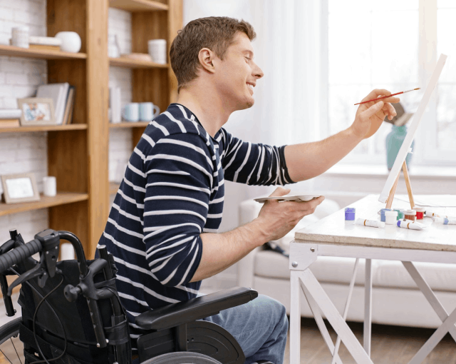 Man in a wheelchair painting at an easel in a bright home environment, illustrating the impact of a stroke and recovery activity.