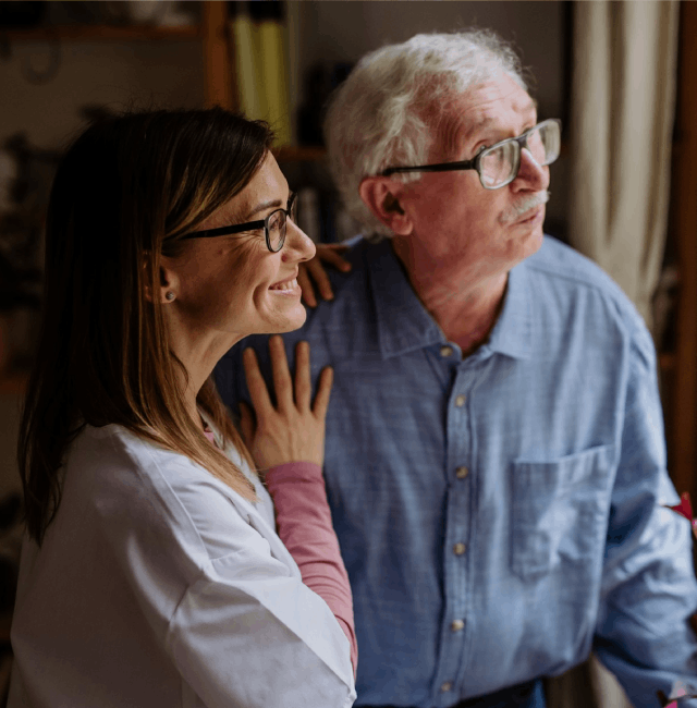 Smiling female caregiver standing beside an older man with glasses, both looking off-camera in a home setting, illustrating the idea that ‘your home is best for stroke care.