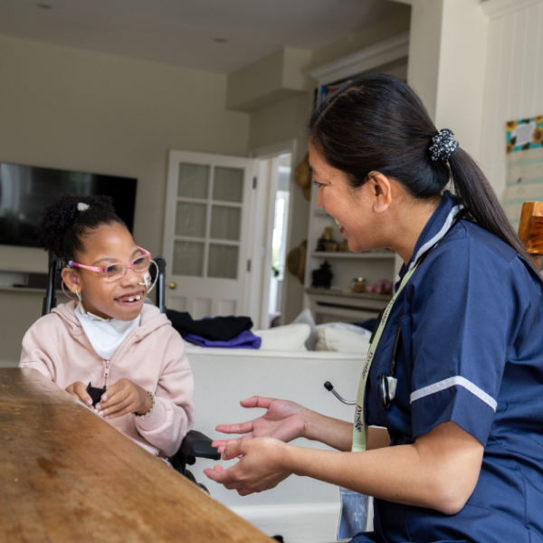 A caregiver in a navy uniform smiling and talking with a young person with cerebral palsy seated at a table, showing supportive at-home care and engagement.