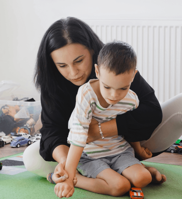 An adult caregiver supporting a young child with cerebral palsy as they play with toy cars on the floor, demonstrating hands-on assistance and engagement.