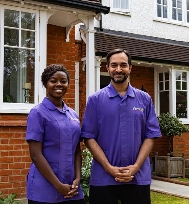 A pair of people wearing Prestige Nursing & Care uniform are positioned in front of a house, appearing cheerful and engaged