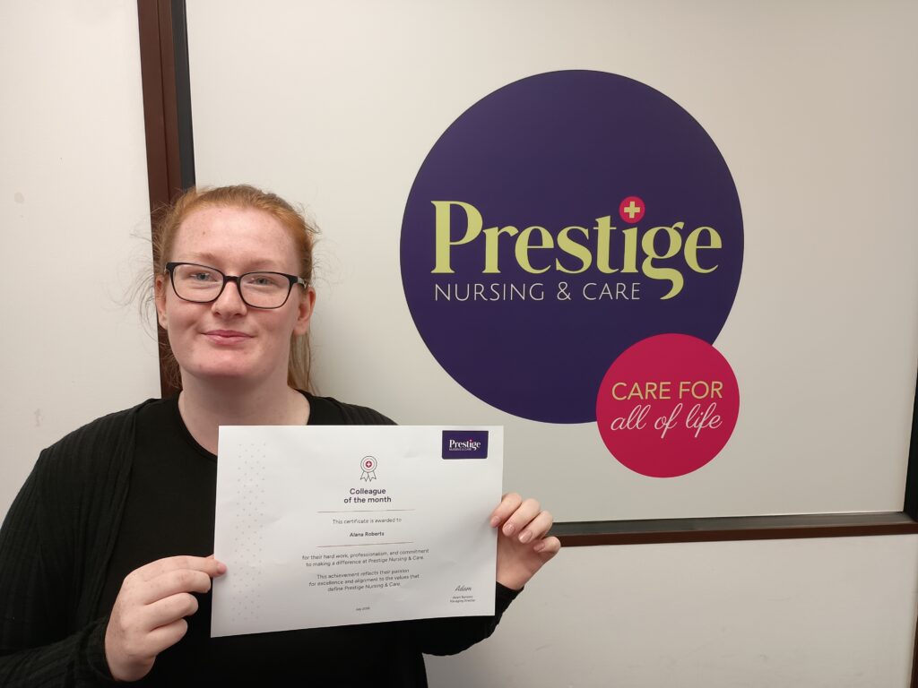 A woman proudly holds a certificate in front of a Prestige Nursing & Care sign, showcasing her achievement and recognition