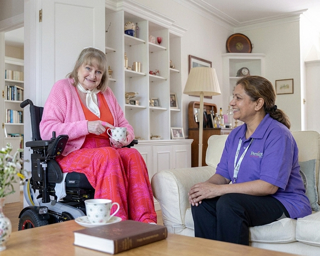 A woman in a wheelchair sits beside another woman in a cozy living room, engaged in conversation
