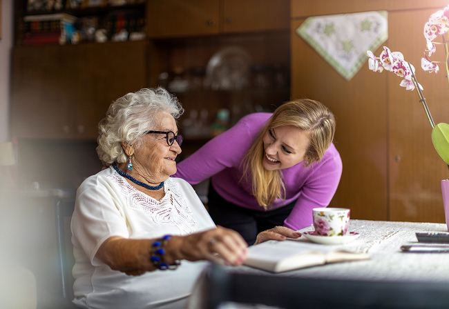 A woman assists an older woman by pointing to text in a book, both engaged in a focused discussion