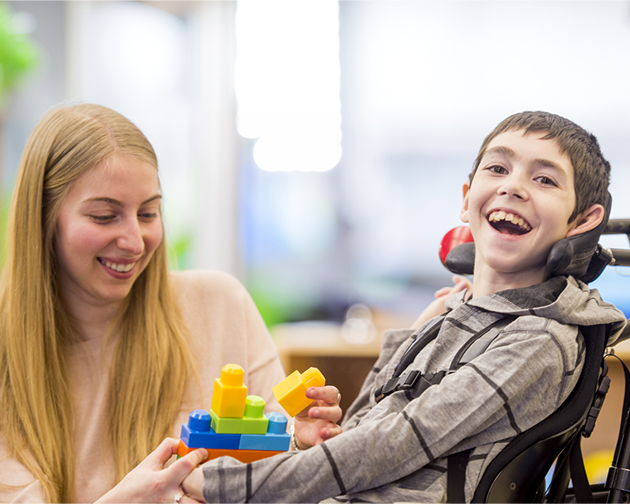 A woman and a boy, both in wheelchairs, share a moment together, smiling in a bright outdoor setting